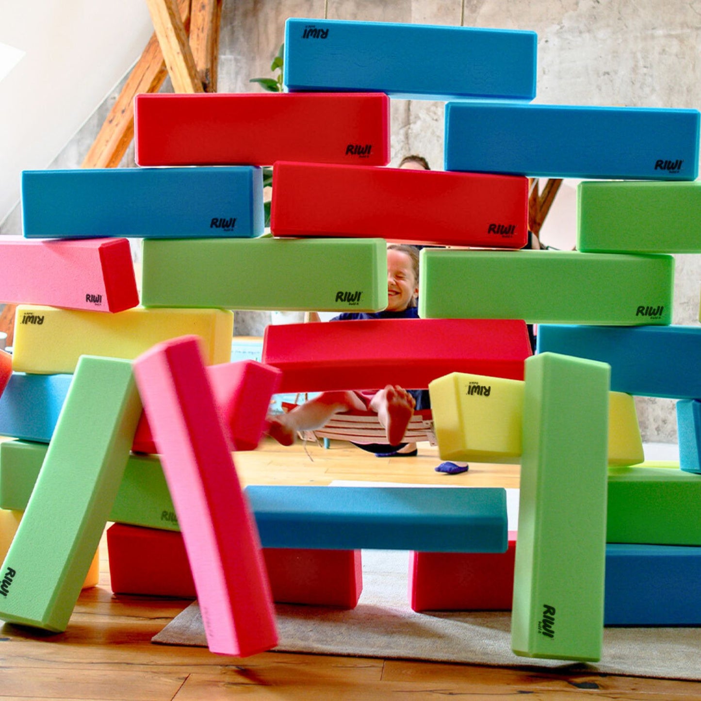 Colorful building blocks stacked in a tower with children playing in the background. RIWI blocks